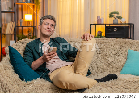 Young man smiles while browsing old family photos on smartphone, enjoying warm nostalgic moment 128077048