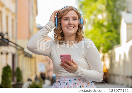 Happy young woman in wireless headphones choosing, listening music in smartphone dancing outdoors 128077151