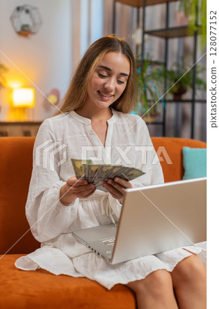 Rich happy young woman counting money cash using laptop calculate domestic bills sitting on sofa 128077152
