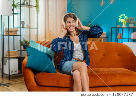 Joyful young woman enjoying listening to music through wireless headphones dancing at home room sofa Joyful young woman enjoying listening to music through wireless headphones dancing at home room sofa 128077177