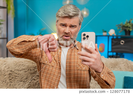 Mature man sitting on sofa with smartphone showing thumbs down, expressing disappointment at home Mature man sitting on sofa with smartphone showing thumbs down, expressing disappointment at home 128077178