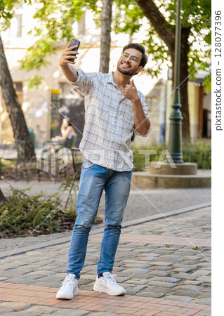 Cheerful Indian man blogger taking selfie on smartphone, talking video call standing on city street 128077396