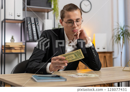 Upset young businessman freelancer counting dollar cash insufficient amount of money at office desk 128077431