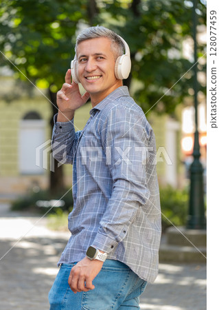 Back view of happy Caucasian male tourist listening to music via headphones walking on city street 128077459
