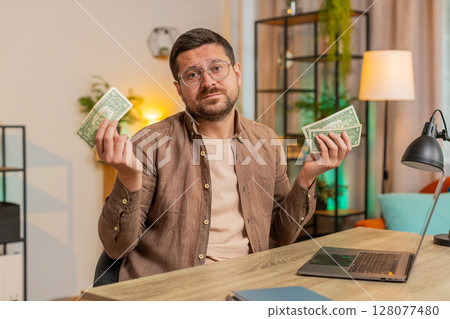 Upset Caucasian male freelancer counting dollar cash insufficient amount of money sitting at table Upset Caucasian male freelancer counting dollar cash insufficient amount of money sitting at table 128077480