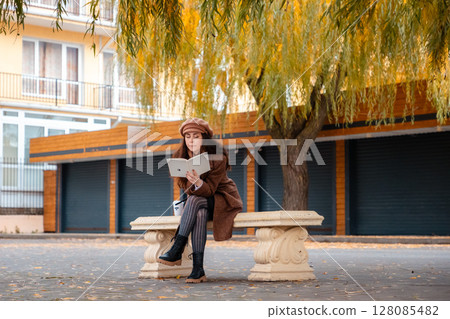 Tranquil weekend scene with a student reading on a city bench in stylish autumn attire. Education and calm outdoor escape. Wide shot Tranquil weekend scene with a student reading on a city bench in stylish autumn attire. Education and calm outdoor escape. Wide shot 128085482