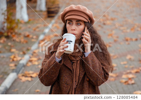 Woman sipping coffee while having a shocked phone call during a walk. Simple urban pleasures and digital connection. Woman sipping coffee while having a shocked phone call during a walk. Simple urban pleasures and digital connection. 128085484