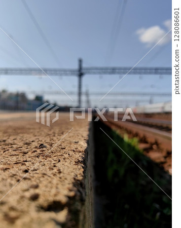 close up view railway station platform with tracks under clear sky and distant trains on sunny day. close up view railway station platform with tracks under clear sky and distant trains on sunny day. 128086601