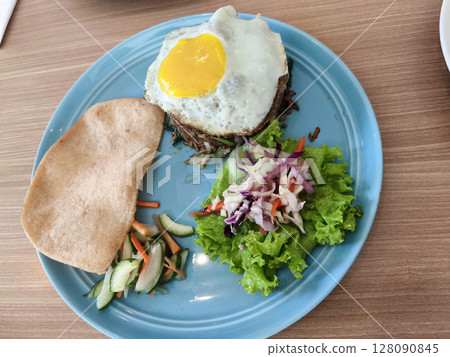 A blue plate holds Indonesian fried rice with a sunny-side-up egg, a large prawn cracker, pickled vegetables, and fresh salad on a wooden table. A blue plate holds Indonesian fried rice with a sunny-side-up egg, a large prawn cracker, pickled vegetables, and fresh salad on a wooden table. 128090845