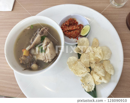 A white plate holds a bowl of Sop Buntut or Indonesian oxtail soup, with a side of chili paste, lime, and crispy emping crackers on a wooden table. A white plate holds a bowl of Sop Buntut or Indonesian oxtail soup, with a side of chili paste, lime, and crispy emping crackers on a wooden table. 128090910