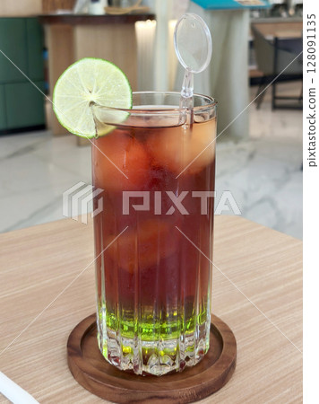A colorful iced tea with a green bottom layer, a lime slice, and a stirrer, sits on a wooden coaster on a wooden table in a cafe in Surabaya, Indonesia. 128091135