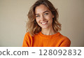 Smiling young woman with wavy hair wearing orange sweater is posing in studio 128092832