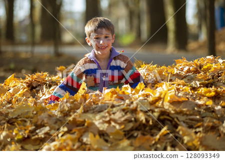 Happy little boy in a pile of yellow leaves in the autumn park. 128093349