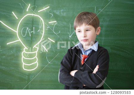 Thinking over an idea. Child student in class. Schoolboy at the green chalkboard with a painted light bulb. Primary school education. Thinking over an idea. Child student in class. Schoolboy at the green chalkboard with a painted light bulb. Primary school education. 128093583