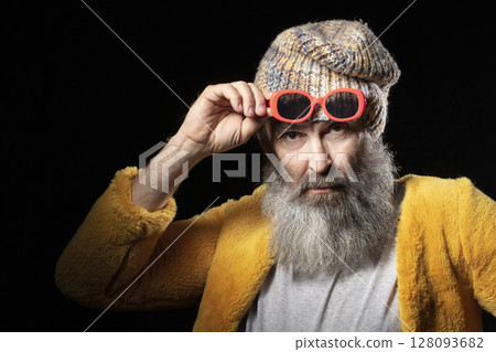 An elderly man with a gray beard, who is wearing a knitted hat and a bright yellow fur coat. Fashionable man on a black background. An elderly man with a gray beard, who is wearing a knitted hat and a bright yellow fur coat. Fashionable man on a black background. 128093682
