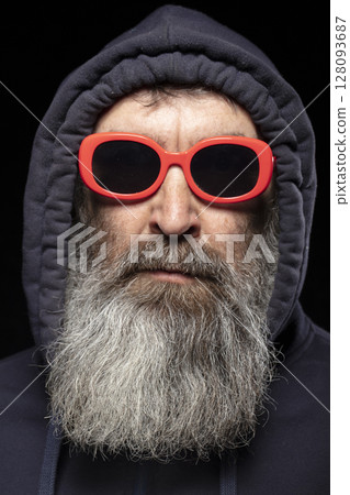 An elderly man with a gray beard in a hood and red glasses on a black background. A man hides something. An elderly man with a gray beard in a hood and red glasses on a black background. A man hides something. 128093687