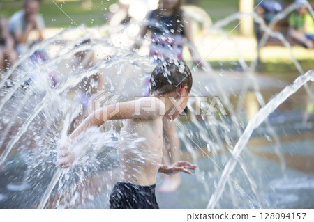 A little boy enjoys the cold waters of a fountain during the heat wave. A little boy enjoys the cold waters of a fountain during the heat wave. 128094157