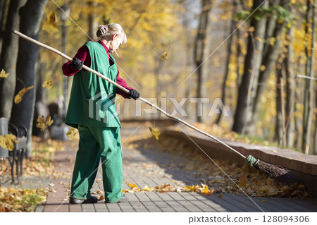 A worker cleans the street with a broom. A janitor cleans the road of fallen leaves in the fall. Utility worker with broom cleaning 128094306