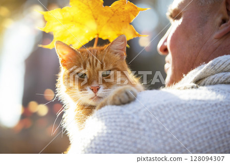 A man is holding a ginger cat and a yellow maple leaf. 128094307