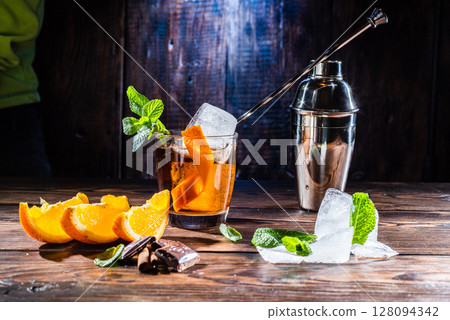 Cocktail with ice and orange. Cocktail spoon in a glass. Orange slices and chocolate in the foreground. Cocktail shaker. 128094342