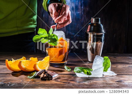 Mixing a cocktail with a bartender's spoon. Bartender mixes a cocktail. Large pieces of ice and mint in a glass. Spoon in hand. 128094343