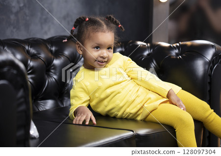 Little beautiful African American girl in a yellow dress sits on a black leather sofa. 128097304