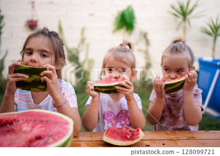 Three girls savoring juicy watermelon slices in sunny garden Three girls savoring juicy watermelon slices in sunny garden 128099721