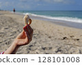 Hand holding seashell on sandy beach with Gulf of Mexico waves and bright blue sky 128101008