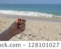 Close up of hand holding shark tooth against sandy Gulf coast background with sea horizon 128101027