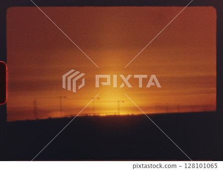 8 mm film - silhouetted electric towers standing against vibrant orange sunset sky, revealing dramatic industrial landscape with warm golden hour light casting serene atmospheric glow across horizon 128101065