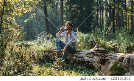 Man enjoying refreshment while sitting on a fallen tree in the forest. 128101802