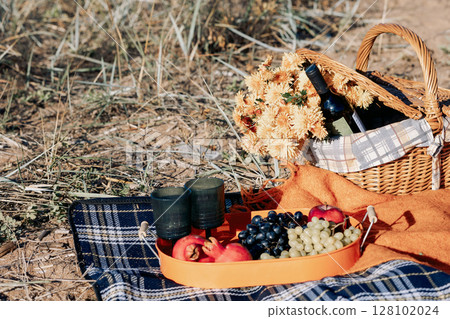 Autumn picnic on beach by sea. Fresh fruits, wine glasses, bottle, grapes, pomegranate near picnic basket on orange blanket. High quality photo 128102024