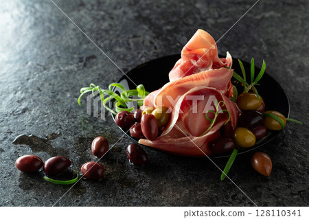Italian prosciutto crudo with olives and rosemary on a black stone table. 128110341