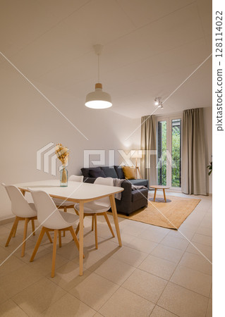 Wide shot of minimalist living and dining area with white dining table, gray sofa, floor lamp and large window 128114022