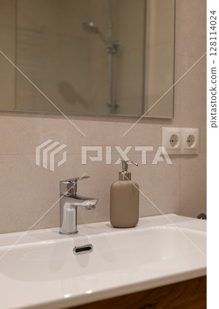 Minimalist bathroom sink area with chrome faucet and beige soap dispenser against neutral tiled wall 128114024