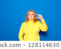 Caucasian woman in her 40s wearing a yellow sweater, smiling and confident, gesturing with her hand, making a small sign with her fingers, looking at the camera. Isolated over blue background. 128114630