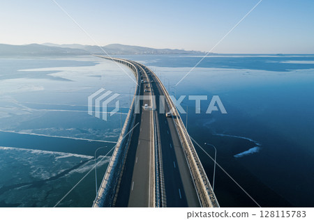 Aerial view of a long road bridge stretching over calm waters at dawn with distant mountains and city skyline Aerial view of a long road bridge stretching over calm waters at dawn with distant mountains and city skyline 128115783