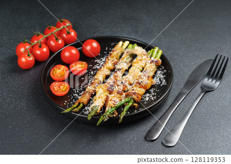 Grilled asparagus wrapped with bacon on black plate with cherry tomatoes on black background Grilled asparagus wrapped with bacon on black plate with cherry tomatoes on black background 128119353
