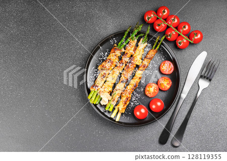 Grilled asparagus wrapped with bacon on black plate with cherry tomatoes on black background Grilled asparagus wrapped with bacon on black plate with cherry tomatoes on black background 128119355