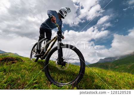 Mountain biker riding down a grassy slope. Enduro mountain bike. MTB against a mountain landscape Mountain biker riding down a grassy slope. Enduro mountain bike. MTB against a mountain landscape 128120528