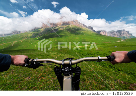First person view of a man's hands holding the handlebars of a bicycle against the backdrop of an epic mountain landscape. Mountain bike concept pov First person view of a man's hands holding the handlebars of a bicycle against the backdrop of an epic mountain landscape. Mountain bike concept pov 128120529