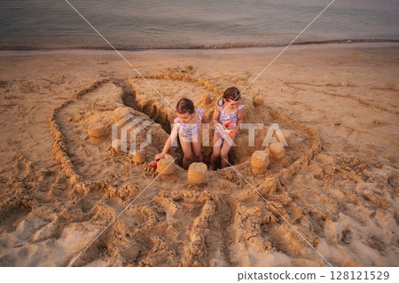 Sisters building a grand sandcastle at sunset on the beach 128121529