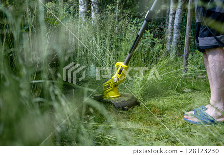 Man trimming overgrown grass with electric string trimmer in summer garden Man trimming overgrown grass with electric string trimmer in summer garden 128123230