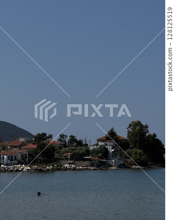 A small Greek village by the water, with red-roofed homes and tree-covered hills, under a clear blue sky 128125519