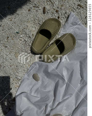 Pair of light green sandals placed on a towel resting over white pebbled sand, captured under direct sunlight on a Greek beach. 128125655