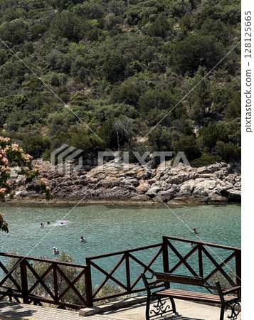 A small rocky cove with clear turquoise water and wooden railing above, perfect for nature, travel, and Greek island visuals. 128125665