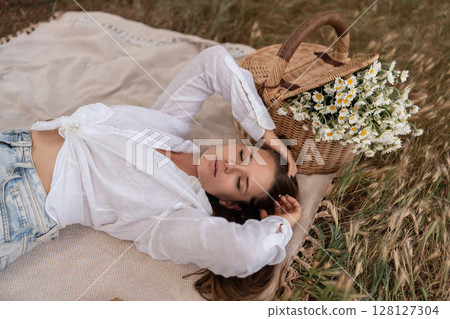 Picnic Daisies Woman: Young woman relaxes on blanket during daytime picnic with flower-filled basket. 128127304