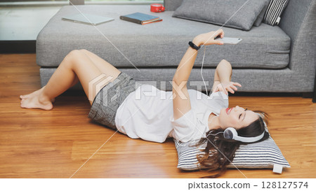 Woman raise her hand greeting her friend via smartphone while lying on the floor at home. Woman raise her hand greeting her friend via smartphone while lying on the floor at home. 128127574