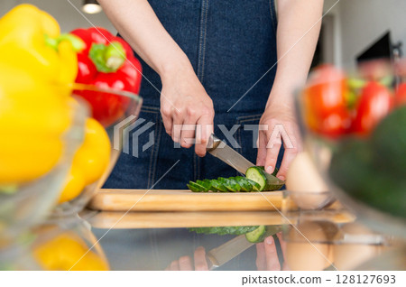 Unrecognizable housewife and her hands cutting green fresh cucumber on wooden cutting board among vegetables and prepared ingredients Unrecognizable housewife and her hands cutting green fresh cucumber on wooden cutting board among vegetables and prepared ingredients 128127693