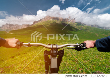 Mountain bike descending from a hill, view from the eyes of a cyclist. First person view of male hands on a mtb bike against the backdrop of rocky mountains wrapped in fluffy clouds Mountain bike descending from a hill, view from the eyes of a cyclist. First person view of male hands on a mtb bike against the backdrop of rocky mountains wrapped in fluffy clouds 128127832